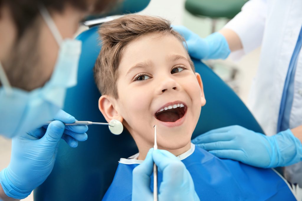 Kid siting in dental chair with mouth wide open and dentist holding tools.