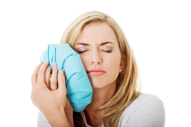 Blonde woman holding ice pack to her mouth with white background.