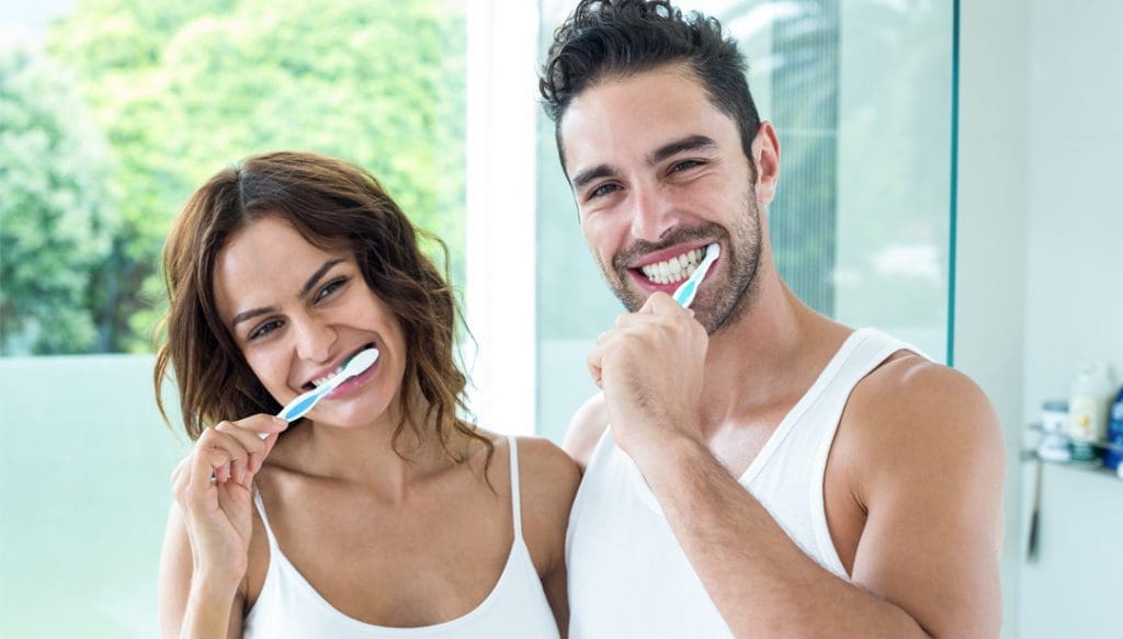 Man and woman side hug as they brush their teeth in the bathroom.