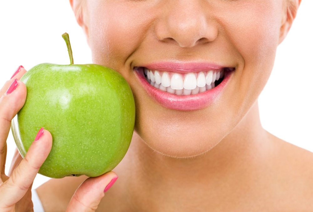 Woman smiling holding green apple to the side of her mouth.