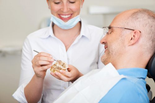 Man sits on dental chair as smiling dentist holds teeth model explaining to him.