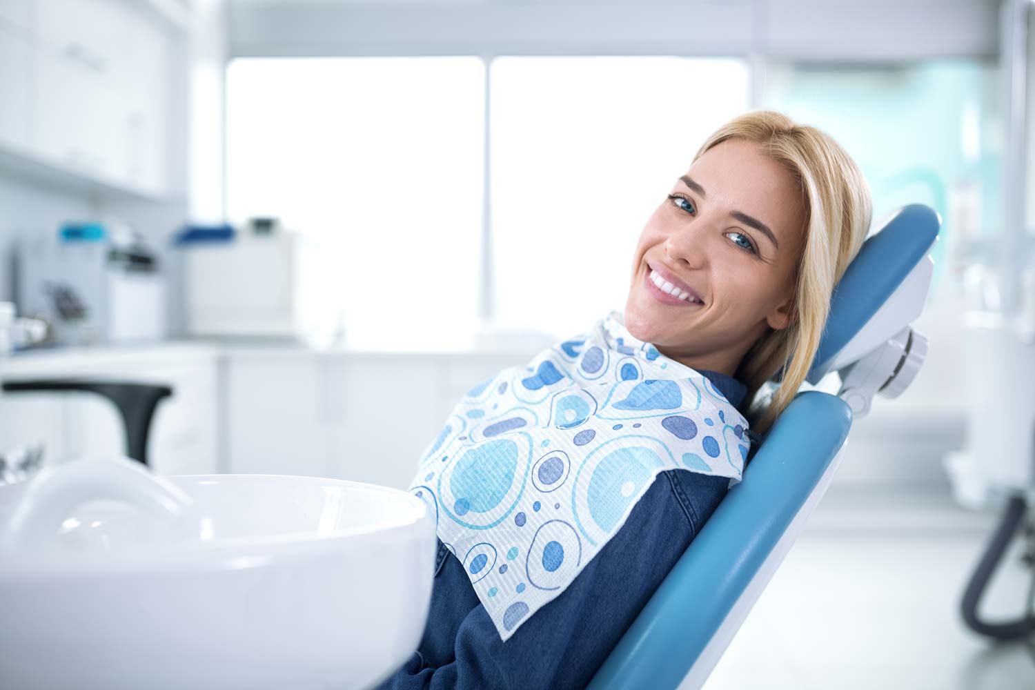 Woman sitting in dentist chair wearing bib with turned head smiles at the camera.