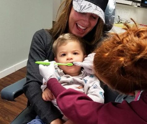 Woman sitting in dental chair holding her young child as a dentist places toothbrush in young Childs mouth.
